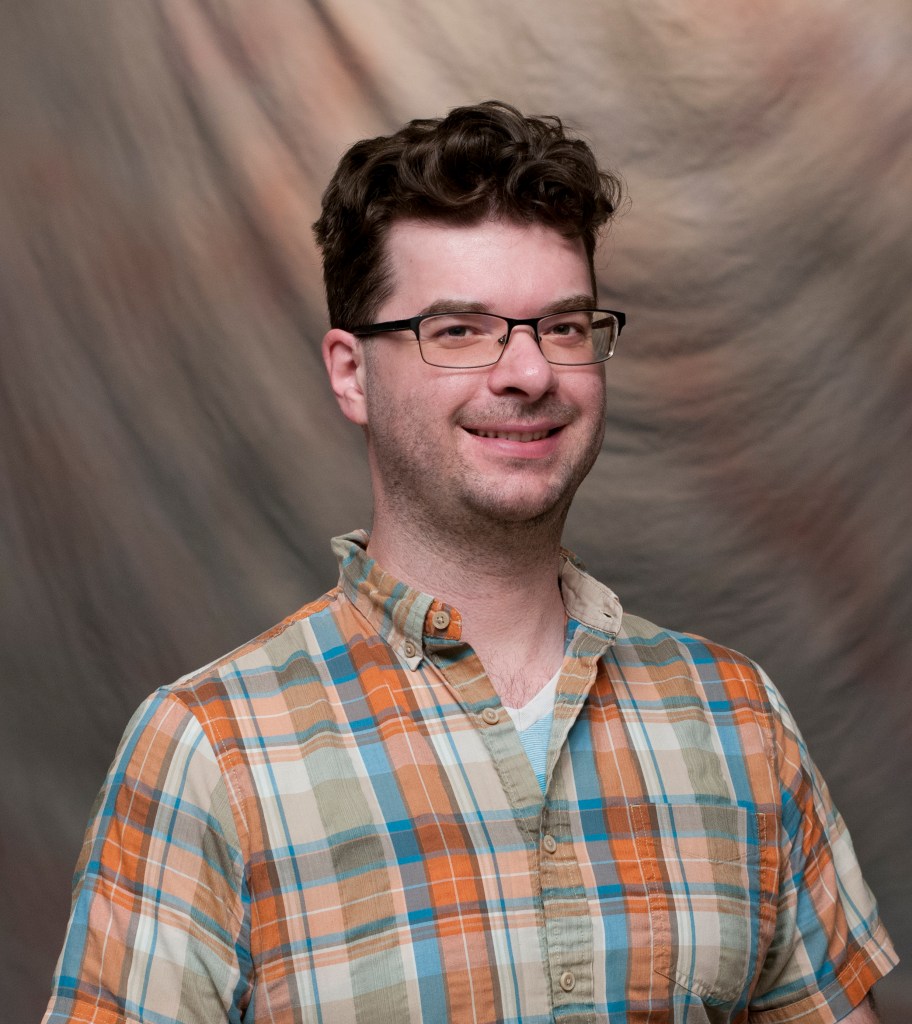 Headshot of Caleb Hutchins smiling. Caleb has dark hair and is wearing glasses and a plaid shirt.