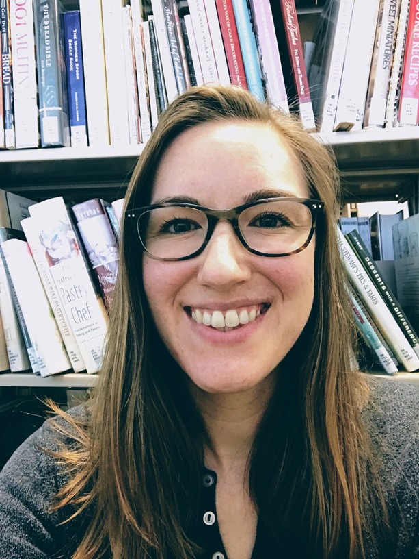 Headshot of Katherine Kelley smiling in front of a shelf of library books. Katherine has long brown hair and is wearing glasses.