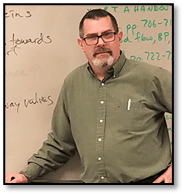 Upper body shot of Stephen Shoemake smiling in front of a whiteboard. Stephen has short dark hair, a graying beard and mustache, and dark eyes.